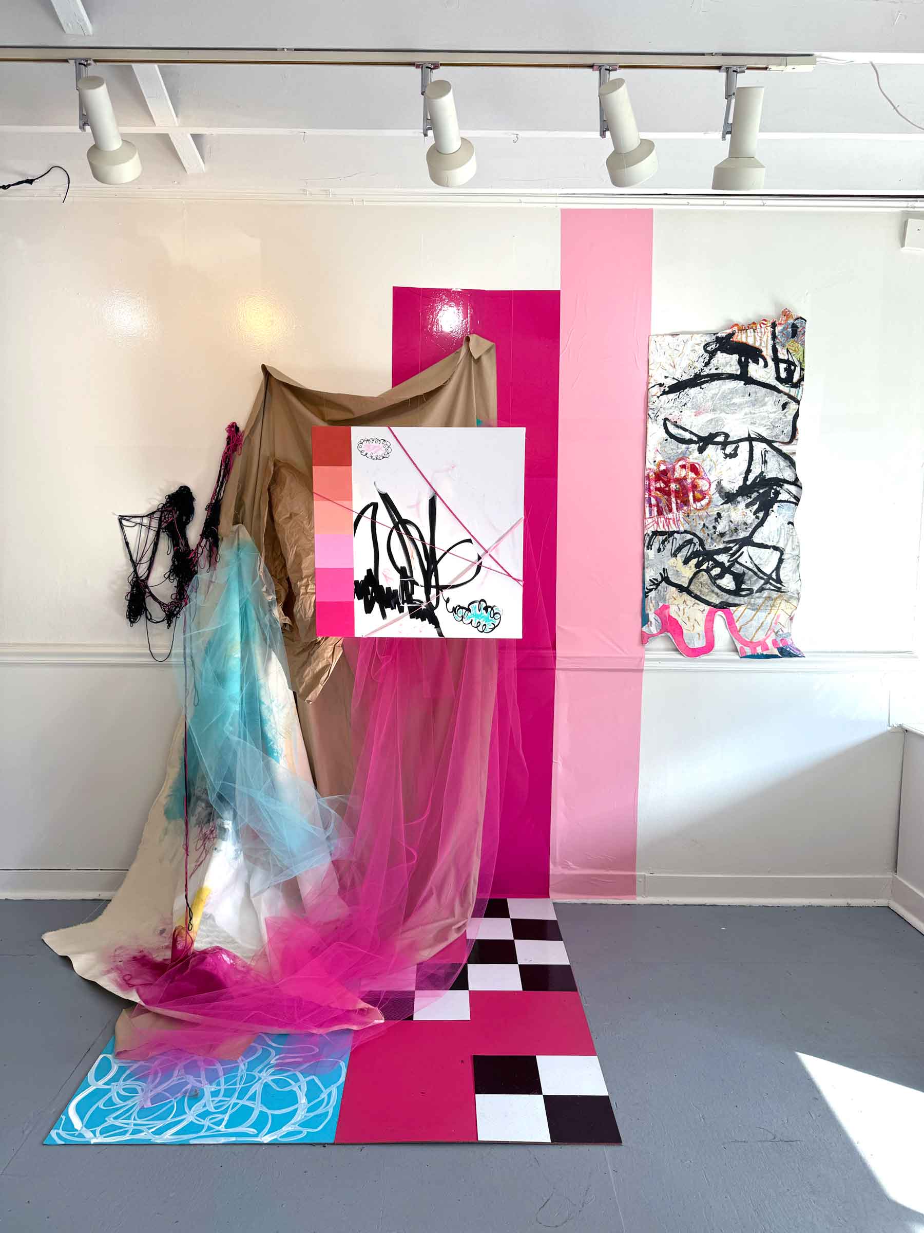 delusion-7.10.836-elizabeth-rennie-3-web Installation mixed media artwork titled ‘Delusion 7.10.836’ by Elizabeth Rennie. The piece features a dynamic assemblage of materials, including draped beige and dyed fabrics, with vibrant splashes of pink and blue. Layers of sheer pink tulle cascade toward the floor, creating a soft, dreamlike quality. A checkerboard pattern of black, white, and red tiles anchors the composition. Abstract painted elements, such as gestural black marks and vibrant pastel gradients, are displayed on a central panel. To the side, textured, crumpled paper and expressive black ink shapes adorn a mounted canvas. The installation is framed by painted pink panels on the wall, giving the piece a vibrant, immersive atmosphere.