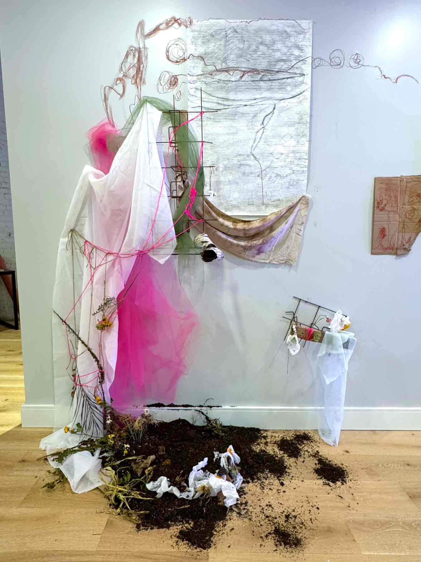 remanents-16.12.04.13.25-2025-elizabeth-rennie Installation mixed media artwork titled ‘Delusion 7.10.836’ by Elizabeth Rennie. The piece features a dynamic assemblage of materials, including draped beige and dyed fabrics, with vibrant splashes of pink and blue. Layers of sheer pink tulle cascade toward the floor, creating a soft, dreamlike quality. A checkerboard pattern of black, white, and red tiles anchors the composition. Abstract painted elements, such as gestural black marks and vibrant pastel gradients, are displayed on a central panel. To the side, textured, crumpled paper and expressive black ink shapes adorn a mounted canvas. The installation is framed by painted pink panels on the wall, giving the piece a vibrant, immersive atmosphere.